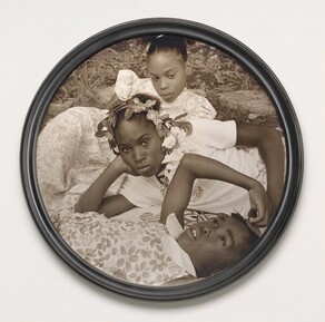 Three young Black girls lie on the grass in this closely cropped, sepia-toned, circular photograph so their faces roughly line up near the center. At the bottom of the composition, a girl, about ten years old, lies on her back and looks up into the sky. Her head, torso, and right arm are visible. She wears a floral-patterned dress and holds her right hand up to the top of her head. The second girl, also about ten years old, reclines on her right side behind the first girl, so she's angled to our left. She props her head in her right hand and looks steadily at us. Her face hovers at the center of the composition. She wears a white t-shirt and a garland encircles her head. The third girl, at the top of the composition, seems to prop her body up on her left elbow. She wears a floral dress and looks down and to our right. Grass and paving rocks fill the space behind her. 