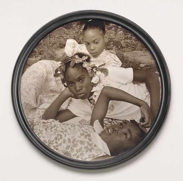 Three young Black girls lie on the grass in this closely cropped, sepia-toned, circular photograph so their faces roughly line up near the center. At the bottom of the composition, one girl lies on her back and looks up into the sky. Her head, torso, and right arm are visible. She wears a floral-patterned dress and holds her right hand up to the top of her head. The second girl reclines on her right side behind the first, so she is angled to our left. She props her head in her right hand and looks steadily at us. Her face hovers at the center of the composition. She wears a white t-shirt and a garland encircles her head. The third girl, at the top of the composition, seems to prop her body up on her left elbow. She wears a floral dress and looks down and to our right. Grass and paving rocks fill the space behind her.