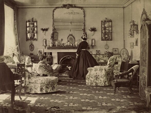 We look toward the far end of a large, well-appointed room at a person reading in an armchair and a woman wearing a long dress standing in front of a fireplace in this horizontal photograph. The image is monochromatic like a black and white photograph but is printed in warm tones of golden and dark browns. Both people have light skin. An expanse of carpet patterned with light flowers against a dark background stretches from where we are to the opposite end of the room. Furniture lines the walls to either side of us, including a desk to our left and a tall cabinet to our right. The scene is lit by ceiling-high windows or doors along the wall to our left. Two armchairs, a chaise, a loveseat, and a sofa, all covered with fabric patterned with dark roses against a light background, are arranged at the far end of the room. A person with short dark hair, wearing a light suit, sits facing away from us, reading a book in an armchair to our left. One hand rests up by the ear we can see, brushing the smooth cheek. The woman standing at the fireplace, facing away from us, wears a white cap on dark hair, and a floor-length, dark dress. The dress has a high, white collar and white cuffs at the end of long sleeves. It is belted at the narrow waist and falls in deep folds, in a wide A-line to the floor. The mantle is lined with a clock and figurines under glass cases. The large, ornately framed mirror fills the height of the wall between the fireplace and the ceiling, which is lined with molding. Other tables set against walls and in front of some of the seating is topped with pictures, figurines, books, plants, and lamps. Cases with small tchotchkes hang on the wall to either side of the large mirror, and other plaques and pictures hang on the walls. The mirror reflects a sliver of the standing woman’s face, showing downcast or closed eyes, a rounded nose, and closed lips. The wall behind us is also reflected in the large mirror, and shows more hanging pictures.