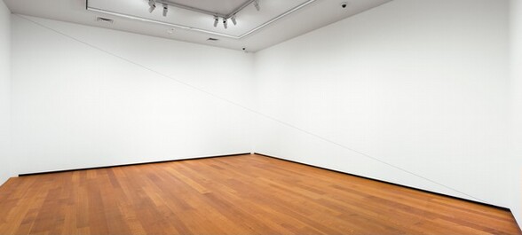 In a room with white walls and hardwood floors, a taut, thin black cord stretches from the upper left to the lower right corner of the room. The cord is barely visible.