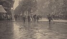 In this black and white horizontal photograph, people and horse-drawn carriages travel along and cross a rain-slicked, urban street. The photograph is roughly divided in half horizontally so the lower half shows a the wet pavement of a wide intersection, momentarily clear of people, where two streets meet in a T intersection. The upper half shows shops and buildings along a tree-lined street at the corner to our left, and along the long, cross street to our right. On the corner, clothing and other wares are displayed outside under an awning , while another display is covered over with a tarp. There are about ten people are on foot crossing the street toward us, while several horse-drawn carriages with large, spoked wheels, driven by caped and top-hatted men, pass through or are parked alongside the curb on the far side of the street. Some of the pedestrians hold up dark umbrellas, while only one man, without an umbrella, crosses the street walking away from us. All of the men wear dark suits and hats. The one woman crossing with the group wears a long skirt and matching thigh-length coat over a contrasting buttoned bodice. She holds up her skirts in one hand and her umbrella in the other. At the far corner, several tall multi-paneled kiosks with domed tops are papered with posters. Across the street is an uninterrupted row of buildings and shops with rows of shuttered windows above. A small triangular slice of sky can be seen between the building at the left and the trees lining the street; its reflection is mirrored on the wet street.