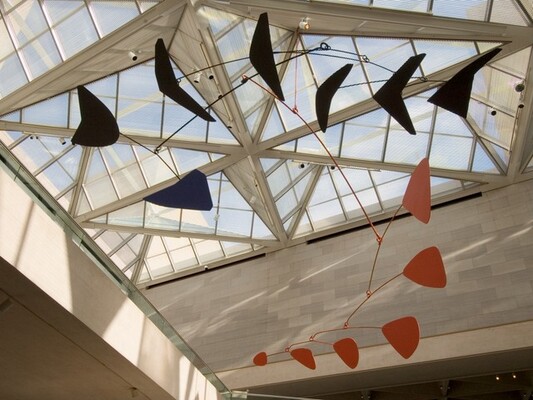Six black, wedgelike shapes, one dark blue arrowhead shape, and six red triangular shapes with rounded corners are connected with thin rods to form this abstract sculpture. In this photograph, we look up at the piece to where it hangs from the glass ceiling of a light-filled interior space with marble walls. The paddle-like shapes are attached to the end of the curved rods, which are linked together like branches. In our view, the black arm curves up across the top of the picture and the red arm curves down into the lower right corner, and to our left. Each red paddle is smaller as they move from the central armature to the end of the branch.