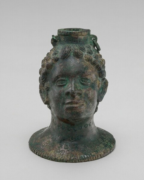 A bronze vessel in the shape of a person’s head and neck faces us with the chin tilted slightly up in this photograph. The surface has aged to form a natural green patina, which is most intense at the inner corners of the lidded eyes. The head has a square jaw, wide nose, closed mouth, and short, stylized curly hair. The pouring spout emerges from the top center of the head. The rim of the spout flares like ears on either side and is pierced with a small circular hole. A ring hangs on our right side, resting against the hair. A row of circles has been incised or punched around the neck of the spout and around the neck of the person, near the chin. Each circle is punched with a dot at its center. Bands of circles decorate the flaring foot of the vessel as well.