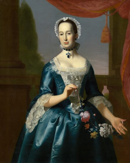 Shown from the knees up, a woman wearing a shimmering teal-blue gown and holding a garland of flowers stands in front of columns and a landscape in this vertical portrait painting. The woman’s body faces us, but she turns her head slightly to our left as she looks at us with dark brown eyes. Her skin is pale, nearly the color white, and she has rosy cheeks. She has an oval face, a long, thin nose, the suggestion of a double chin, and her peach-colored lips are closed. Her brown hair is pulled back under a lacy cap, and she wears dime-sized sapphire earrings rimmed with gleaming silver or diamonds. A pendant with three round sapphires and a larger, teardrop-shaped gem hangs from a choker made of thumbprint-sized sapphires. The edges of a white bow, presumably tying her hair, curl to either side of her neck. Her tight-fitting bodice has a square, lace-edged neckline and white ribbons tied down the front. Long, lacy cuffs fall from elbow-length sleeves, and the full skirt falls in vertical folds from the cinched waist. The teal fabric has a sheen, suggesting silk or satin. A pink rose as well as white, yellow, orange, and blue flowers are tied to a pink ribbon to create a garland, which she holds with both hands. Tan-colored stone columns lining the painting to either side behind her are connected by a balustrade, all of which frames a view out to a tall, light green tree and a blue sky touched with pink along the horizon. A coral-pink curtain hangs in the top right corner.