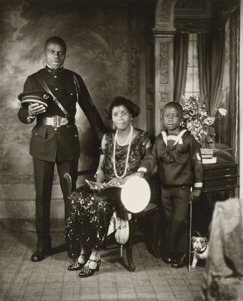 A Black woman, young man, and young boy pose against a painted backdrop in this vertical photograph. The image is monochromatic like a black and white photograph but is printed in warm tones of golden and dark browns. The man and woman have slight smiles, and all three look at us. At the center, the slender woman sits on a wooden chair with her body angled slightly to our left, though she turns her head to us. Her skin tone is a little lighter than that of the man and boy, and her hair is styled in an ear-length bob. Her scoop-neck, sleeveless, ankle-length dress shimmers as if covered with beads or sequins. She wears round glasses, dangling earrings with pearls, and short and long strands of pearls. Light catches two rings on her left hand and a bangle bracelet encircles each arm. Her shiny, high-heeled shoes have a lattice-like pattern cut into the tops around the T-straps. She holds an object, perhaps with feathers, in her lap. To our left of the woman, the young, cleanshaven man stands facing us with his weight equally balanced on both feet, positioned slightly apart. He rests his left hand, on our right, on the back of the woman’s chair and holds his flat-topped, brimmed hat in the crook of his other elbow. His dark, crisply pressed uniform has a snug fitting jacket with a high collar, shiny buttons in a row down the front, and a braided cord looped over his left shoulder and under his arm. The jacket is cinched with a wide belt and a thin leather band runs diagonally across his chest. To our right of the woman, the boy stands with one ankle crossed in front of the other and his right arm, on our left, resting on the arm of the woman’s chair. In that hand, the top of his hat is a bright white oval. He rests his other hand near the top of a child-sized cane, close to his body. His hair is cut very short. The boy’s dark sailor’s suit has white stripes around the neck and at the cuffs. A white bulldog puppy with darker spots around its eyes stands at the boy’s feet, partially hidden by fabric that hangs off a piece of furniture in the lower right. A wooden table behind the boy holds a stack of a few books and a bouquet of wilted, light colored flowers. The background behind the people has a painted or wallpapered section to our left and an arch leading to a curtained window to our right. Parts of the photograph are noticeably out of focus, particularly the background.
