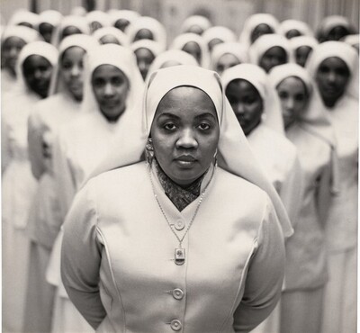 Shown from the waist up, a Black woman, Ethel Shariff, wears a nun’s habit and light-colored clothing as she stands in front of about two dozen more Black nuns in this square black and white photograph. Ethel is in sharp focus at the center of the picture. She looks steadily at us with wide-set eyes, a rounded nose, and full lips in her oval face. The habit covers her head and falls beyond her shoulders. A scarf is tucked into the high neck of her white garment, which buttons up the front. She wears chunky earrings and a glimmering gold necklace with a small box-shaped pendant. She and the other women stand with their hands clasped behind their backs. Those behind Ethel stand in formation, creating a loose V. They are out of focus, their faces a variety of tones.