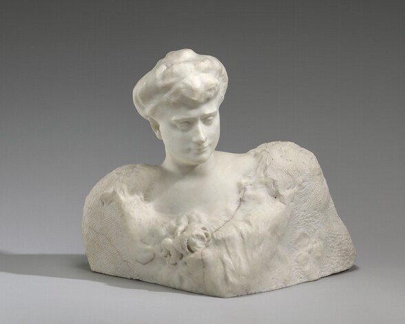 Carved from white marble, the head and chest of a woman emerge from a base of rougher stone in this free-standing sculpture. In this photograph, the woman leans slightly to our left and her head tips down that way, as she looks off to our right. Her hair is loosely pulled up into a bun on top of her head. She has a straight nose, thin lips, and a square jaw. A bumpy area at the front of her low-cut bodice suggests a floral corsage. Her neck, the top of her shoulders, and her chest are polished smooth, but below the scooped neckline, the stone turns rough and parallel chisel marks are visible. Two dark veins in the marble run through the sculpture near each shoulder.