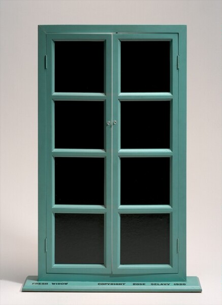 This free-standing sculpture is a French window with a teal-colored frame and black, leather-lined glass panes standing on a flat, shallow base. Each door is made up of four, vertically stacked, equally sized panes. The doors are hinged on a narrow frame and open with small, clear knobs placed next to the second pane down from the top. The doors are slightly ajar. Black writing in capital block letters on the top surface of the base is legible in this photograph. It reads, “FRESH WIDOW COPYRIGHT ROSE SELAVY 1920.”