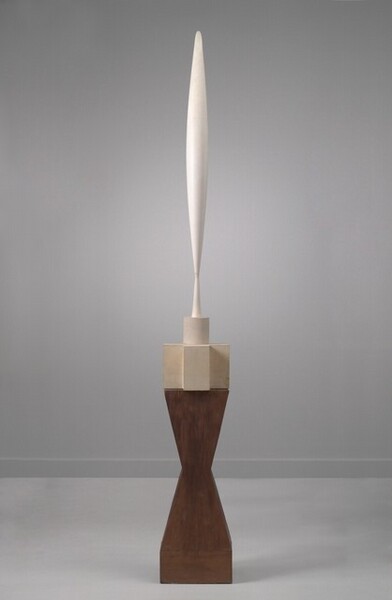 The top half of this modern, abstract sculpture is made up of a vertical, elongated form that swells gently at the center and tapers to a point at either end, like the point of an elephant’s tusk. Near the bottom point, the form flares out slightly to make a tall, conical foot. The marble section sits atop a base made of three stacked pieces that together are about the same height as the swelling form. First is a short cylinder that sits on the center of a piece that would look like a plus sign if viewed from above. Both the cylinder and plus-sign are carved from cream-colored marble, so are a little darker than the whiter, swelling form above. The largest, tallest piece is the dark brown wooden base below, which is carved in the shape of an angular hourglass.