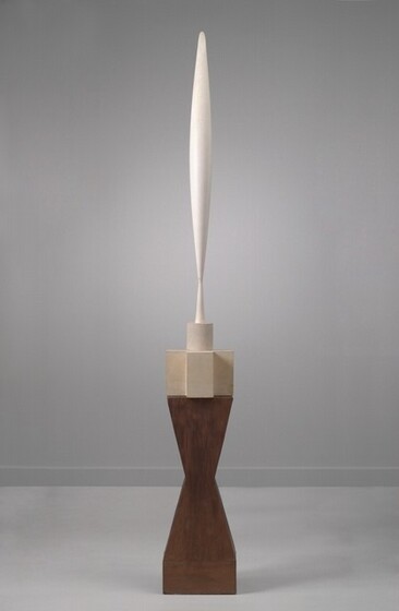 The top half of this abstract sculpture is made up of a vertical, elongated form carved from white marble that swells gently at the center and tapers to a point at either end. Near the bottom point, the form flares out slightly to make a tall, conical foot. This sits atop a base made of three stacked pieces that together are about the same height as the swelling form. First, just below the elongated form, a short cylinder sits on the center of a piece that would look like a plus sign if viewed from above. Both the cylinder and plus-sign are carved from cream-colored stone, so are a little darker than the whiter, swelling form above. The tall, dark brown wooden base below is carved in a shape like an angular hourglass.