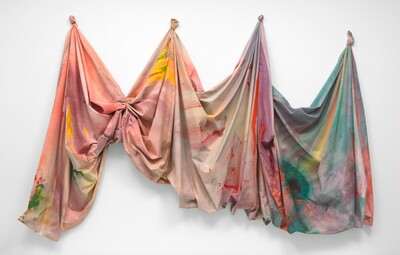 This painted canvas hangs on the wall loosely from four gathered peaks—one peak on each end to the left and right, and two peaks evenly spaced in between. The fabric is tightly wrapped with a leather cord into a fist-like form to create each peak, except for the right-most peak, where the fabric is knotted. The canvas is stained with large areas of soft color that largely meld together, with mostly pink, peach, and yellow to the left that transitions to violet, turquoise, and sky blue to the right. Hard-edged, vivid orange streaks break through the blues and greens to the right.