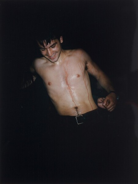 A smiling, bare-chested, pale-skinned man dripping with blood emerges from a pitch-black background in this vertical photograph. He is clean-shaven and has short, dark hair, thick brows, and pointed features. Pale blood trickles over his shoulders and down his sweaty torso as he leans to our left. One hand emerges from the shadows to hold his left hand, on our right. A silver belt buckle gleams against the black pants, which disappear into the inky background.