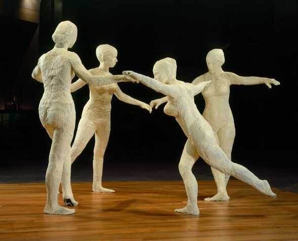 Four ivory-white, free-standing, nude women dance in a circle on a wooden surface against a deeply shadowed background. In this photograph, the two women on the far side of the circle face us, one to our left turns away from us, and the one closest to us faces our left in profile. The one facing our left leans forward with her arms spread wide at shoulder height. Her near leg extends back, and the other leg is bent slightly at the knee. The woman to our left has her back to us. Her right arm reaches out to her right, and she steps forward onto the ball of her left foot. The third woman stands beyond and between the first two. She is angled to our right with her arms spread outward and her right leg, on our left, stepping back. The final woman is on the right and faces us with her head bowed down and to our left. She spreads her arms out just below shoulder-height, and her feet are turned in opposite directions.