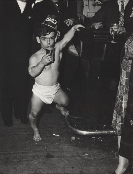 This vertical, black and white photograph focuses on a dark-haired, barefoot man of short stature wearing a diaper and party hat, which bears the year, 1943. His left hand, to our right, reaches overhead and rests on a bar. His left foot perches on the footrail, and he holds a full glass to his pursed lips. He is surrounded by a black field, which eventually becomes evident as people wearing dark suits, their heads cropped by the top edge of the image.