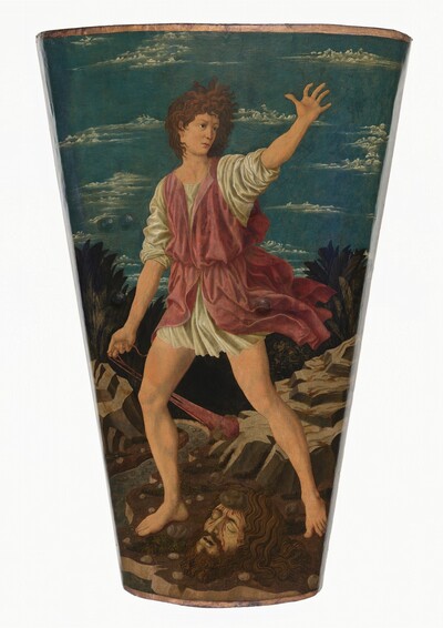 This shield-shaped painting shows a young man with pale, peachy skin and curling brown hair standing astride the severed head of a man with dark hair and tanned skin. The painting is wider at the top and tapers like a pint glass to a narrower base. The young man, David, holds the end of long red slingshot with his right hand. The slingshot is weighted by a rock and swings behind his body. His sleeveless rust-red garment is cinched at his waist over a long white shirt. Both garments skim David’s thighs and his legs and feet are bare. Long, curling brown hair frames the ashen and bloodied upturned face of the head at David’s feet. A palm-sized stone is embedded in that forehead. The landscape background is made up of gray boulders, a meandering river, and low palm-like trees. Frothy white clouds sweep across an azure-blue sky.