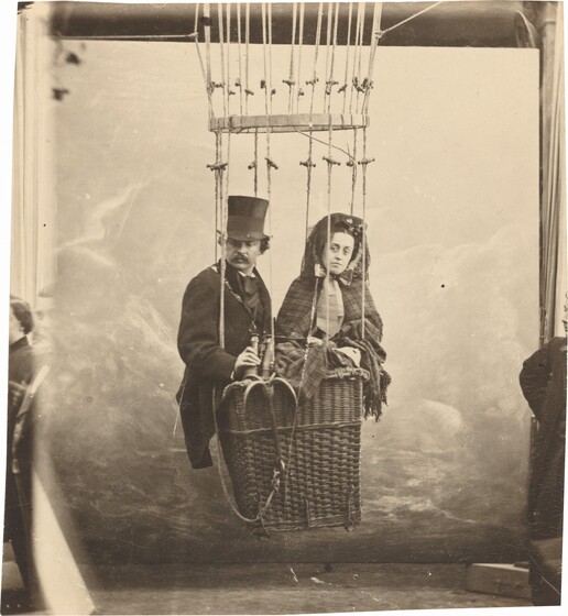 A mustachioed man and a woman, both with light skin, crowd in the basket of a hot air balloon in this vertical, sepia-toned photograph. To our left, the man wears a tall top hat and a dark coat and cravat. With his body angled slightly to our right, he holds a pair of binoculars at his waist as he looks down and off to the side to our left. On our right, the woman wears a plaid patterned shawl that wraps around her body and seems to cover her head. A bow tied under her chin is a little wider than her face, and the wide ends of the ribbons reach far down her chest. She looks at us. The profile and torso of a person is cropped by the right edge of the photograph. Building on that, we eventually realize that the basket hangs a few feet above the floor in front of what must be a painted backdrop in a studio. The left edge is blurred and a sliver showing other people, or perhaps another exposure, is visible.