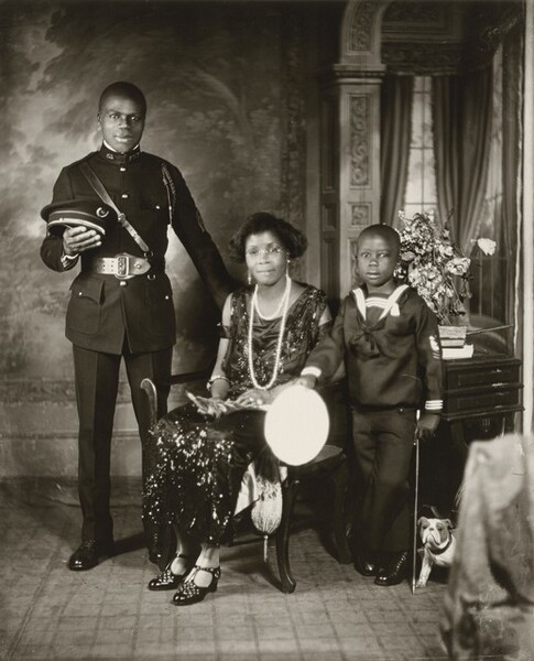 A Black woman, young man, and young boy pose against a painted backdrop in this vertical photograph. The image is monochromatic like a black and white photograph but is printed in warm tones of golden and dark browns. The man and woman have slight smiles, and all three look at us. At the center, the slender woman sits on a wooden chair with her body angled slightly to our left, though she turns her head to us. Her skin tone is a little lighter than that of the man and boy, and her hair is styled in an ear-length bob. Her scoop-neck, sleeveless, ankle-length dress shimmers as if covered with beads or sequins. She wears round glasses, dangling earrings with pearls, and short and long strands of pearls. Light catches two rings on her left hand and a bangle bracelet encircles each arm. Her shiny, high-heeled shoes have a lattice-like pattern cut into the tops around the T-straps. She holds an object, perhaps with feathers, in her lap. To our left of the woman, the young, cleanshaven man stands facing us with his weight equally balanced on both feet, positioned slightly apart. He rests his left hand, on our right, on the back of the woman’s chair and holds his flat-topped, brimmed hat in the crook of his other elbow. His dark, crisply pressed uniform has a snug fitting jacket with a high collar, shiny buttons in a row down the front, and a braided cord looped over his left shoulder and under his arm. The jacket is cinched with a wide belt and a thin leather band runs diagonally across his chest. To our right of the woman, the boy stands with one ankle crossed in front of the other and his right arm, on our left, resting on the arm of the woman’s chair. In that hand, the top of his hat is a bright white oval. He rests his other hand near the top of a child-sized cane, close to his body. His hair is cut very short. The boy’s dark sailor’s suit has white stripes around the neck and at the cuffs. A white bulldog puppy with darker spots around its eyes stands at the boy’s feet, partially hidden by fabric that hangs off a piece of furniture in the lower right. A wooden table behind the boy holds a stack of a few books and a bouquet of wilted, light colored flowers. The background behind the people has a painted or wallpapered section to our left and an arch leading to a curtained window to our right. Parts of the photograph are noticeably out of focus, particularly the background.