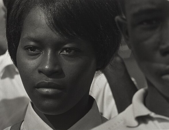 The face of a young Black woman fills most of this horizontal black and white photograph. The top of her head is cropped. She has straight black hair, which sweeps down in bangs across her forehead, and her hair is either cut short or pulled back. Her lips are closed and she looks steadily with dark eyes off into the distance to our left. Only the white or light-colored collar and very top of her right shoulder, on our left, are visible, though a dark narrow strap over her shoulder near her neck suggests a purse or backpack. She is positioned slightly to our left and to our right, the left half of a young Black man’s face is cropped by the right edge of the photograph. He stands closer to us, in front of the woman’s shoulder. His face is out of focus and he looks directly at the camera. More people fill the background between the woman and man but specific features are indistinguishable.