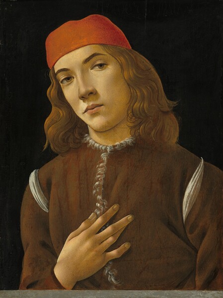 This vertical portrait shows the head, shoulders, and chest of a young man with smooth, white skin and shoulder-length, dark blond hair positioned behind a narrow gray, stone ledge and against a black background. He tilts his head to our left and his eyes cut to our right. His left eyebrow, on our right, is slightly arched over brown eyes. He has a long, straight nose, pale pink lips, and his hair falls down the sides of his face from beneath a crimson, flat-topped cap. He wears a brown garment with white linen peeking through at the shoulder seams. The neck and seam down the front are lined with white, perhaps fur. He holds his right hand to his chest with his middle finger overlapping his ring finger to make a W shape.