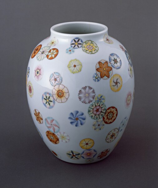 This white porcelain vase is almost egg-shaped, wider at the top and slightly tapered toward the bottom. The round opening at the top center has a low rim. The vase is decorated with circular, stylized flowers with almost geometric patterns made up of butter yellow, peach, ivory, pumpkin orange, and shades of blue. The blossoms seem randomly scattered so some cluster and overlap while others float alone.