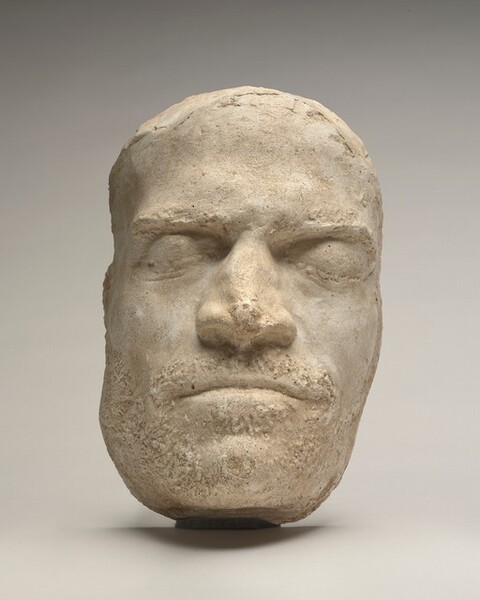 This plaster cast captures a man’s long face, bumped, broad nose, and thin-set lips. The man’s deep-set eyes are closed under a low brow. He has high cheekbones and a square jaw. Texture around his mouth and on his jaw suggests stubble or facial hair.