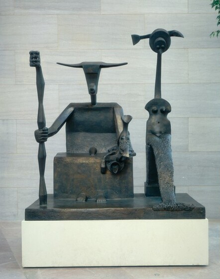 Two abstracted creatures, one resembling a goat holding a staff and the other a stylized mermaid, sit facing us on blocks on a rectangular base in this bronze sculpture. The head of the goat creature, to our left in this photograph, has wide horns to either side of a ridge suggesting a heavy brow. The triangular head tapers down to a ring that suggests the muzzle of the goat. The body is made up of broad geometric shapes. There is a stylized phallus where the body meets the block we read as a lap, and two rounded forms suggestive of human feet protrude from the base of that block. The creature reaches out with his right arm, to our left, to wrap a large, human fist around the staff. The staff swells and narrows in angular hourglass shapes and is topped by what could be a rectangular, abstracted face with two eye holes, a flattened nose, and a slit for a mouth. The goat’s opposite hand rests near that knee, and in it he holds a creature about the height of the goat’s torso. The creature has a scooped, cup-like form where the head would be, a long neck, and two mounds reminiscent of breasts above a textured fish’s tail. Below, as if affixed to the front of the goat’s lap, a disk-like face, like an emoji, has two rings for eyes and a tongue protruding from a round mouth. Other forms over the forehead and one next to the face read as flattened hands or claws. To our right, the second, taller creature has a head made up of a disk with two mounds for eyes and a beak stacked in front of a second, larger disk. A form like an abstracted fish or arrow moves through or behind her head. The creature has a long, slender neck and an armless torso, shaped roughly like the body of a violin. The torso has rounded shoulders, two mounds to suggest breasts, and a hole for a belly button. Her textured fish’s tail curves down so the fin falls slightly over the front of the base on which they sit. The bronze surface of the sculpture varies from dark brown to faint gold where the light catches it. The sculpture and bronze base sit on a white platform. The floor beneath is pale pink, shiny marble and the wall behind is made up of stone panels streaked with gray.