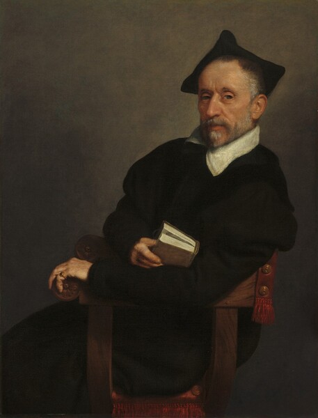 Shown from the knees up, a light-skinned man wearing a loose, ink-black garment with a white collar leans back in a wooden chair in this vertical portrait painting. His body faces our left almost in profile, but he turns his head to look at us from the corners of his light hazel-brown eyes, the eyebrow closer to us slightly cocked. Crow’s feet line the corners of his hooded eyes. He is lit from our right so the side of his face to our left is in shadow. His slightly hollow cheeks are lined between his nose and mouth with wrinkles. His silvery-gray beard is trimmed close to his angled jawline and pointed chin. His gray hair is closely cropped hair under a velvety-black, three-pointed cap. The white collar is trimmed with stripes and a row of delicate dots along the edge. The man leans heavily against his left arm, closer to us, which is stretched along the arm of the chair to nearly span the width of the painting. That hand curls around the terminal of the chair, which is carved with a stylized flower in a disk. He wears a gold ring with a dark blue stone on the pinky of that hand. He marks the spot in a closed book with his other hand, and rests the book in the crook of the elbow draped along the arm of the chair. The book he holds has a sand-brown cover, and is about the size of a thick paperback novel. The crimson-red lining along the back of the chair, to our right, and the cushion on which the man sits is trimmed with rows of fringe of the same color. The background behind the man is deep, sable brown in the corners and lightens to tan around the man’s head.
